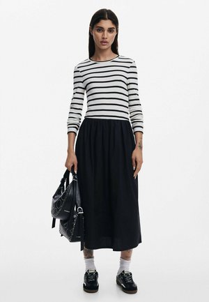 Zwart-witte gestreepte lange mouwen top, zwarte midi rok, gecombineerd met een zwarte tas en donkere sneakers. Opvallende tattoo's op de polsen.