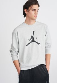 Grauer Hoodie aus Baumwollmischung mit einem schwarzen Jordan-Logo auf der Vorderseite. Das Modell trägt ihn mit den Händen in den Taschen, kombiniert mit einer dunklen Hose.