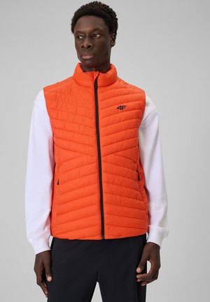 Homme portant un gilet matelassé orange vif sur une chemise blanche à manches longues, debout devant un fond gris clair, regardant sur le côté.