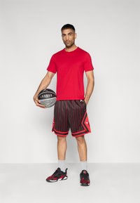 Κόκκινο κοντομάνικο μπλουζάκι με μαύρο λογότυπο, σορτς με μαύρες και κόκκινες λεπτές ρίγες, γκρι αθλητικές κάλτσες και μαύρα/κόκκινα αθλητικά παπούτσια. Κρατάει ένα μπάσκετ.