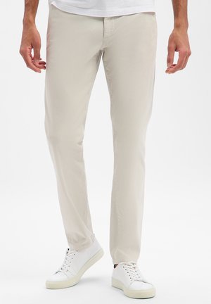 Homme portant un pantalon beige à jambes droites et des baskets blanches, debout avec les mains détendues le long du corps sur un fond blanc.