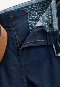 Pantalon bleu marine avec fermeture éclair partiellement ouverte, bouton ouvert et ceinture à motif floral visible, associé à une ceinture marron.