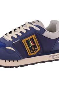 Sneaker blu navy con dettagli grigio chiaro, tessuto testurizzato e una prominente toppa gialla con logo e design tricolore. Suola spessa beige.