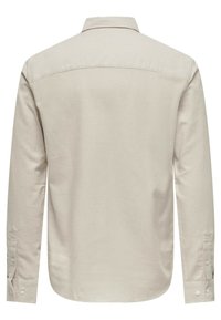 Camisa de manga larga beige con cuello, puños abotonados y una textura de algodón suave. Dobladillo ligeramente curvado en la parte inferior, sin gráficos visibles.