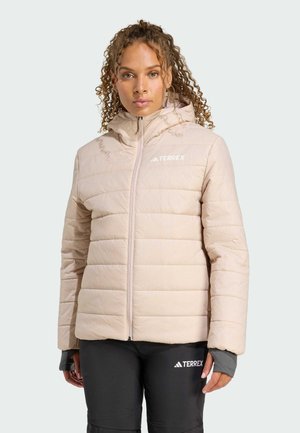 Een persoon met krullend haar draagt een lichtbeige pufferjack en donkere broek, en toont casual buitenschoeisel voor een neutrale achtergrond.