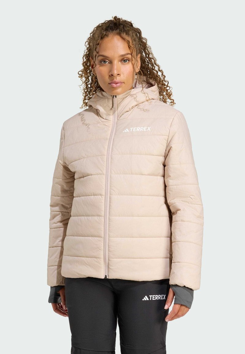Eine Person mit lockigem Haar trägt eine hellbeige Steppjacke und dunkle Hosen, die lässige Outdoor-Bekleidung vor einem neutralen Hintergrund präsentiert.