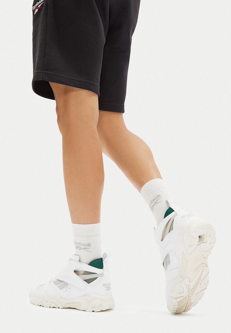 Białe buty sportowe o masywnym designie, z panelami z siateczki i zielonymi akcentami, zestawione z czarnymi szortami i białymi skarpetami.