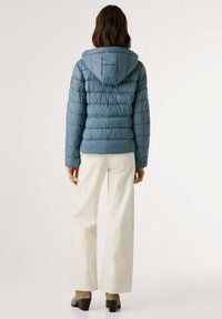 Vrouw in een blauwe gewatteerde hoodie-jack, met witte wijdvallende broek en hakken, staande met haar rug naar de camera op een witte achtergrond.