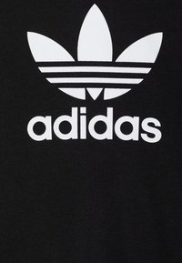 Juodas medvilnės marškinėliai su dideliu baltu Adidas logotipu, virš kurio yra trys lapo formos ir po logotipu trys horizontalios juostos.