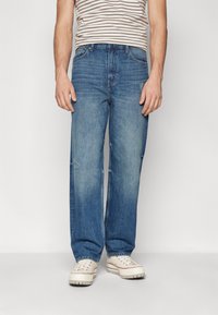 Jeans din denim de culoare albastru deschis, cu o croială lejeră, având zone decolorate și ușoare deteriorări. Asortate cu adidași de culoare crem și o cămașă în dungi.