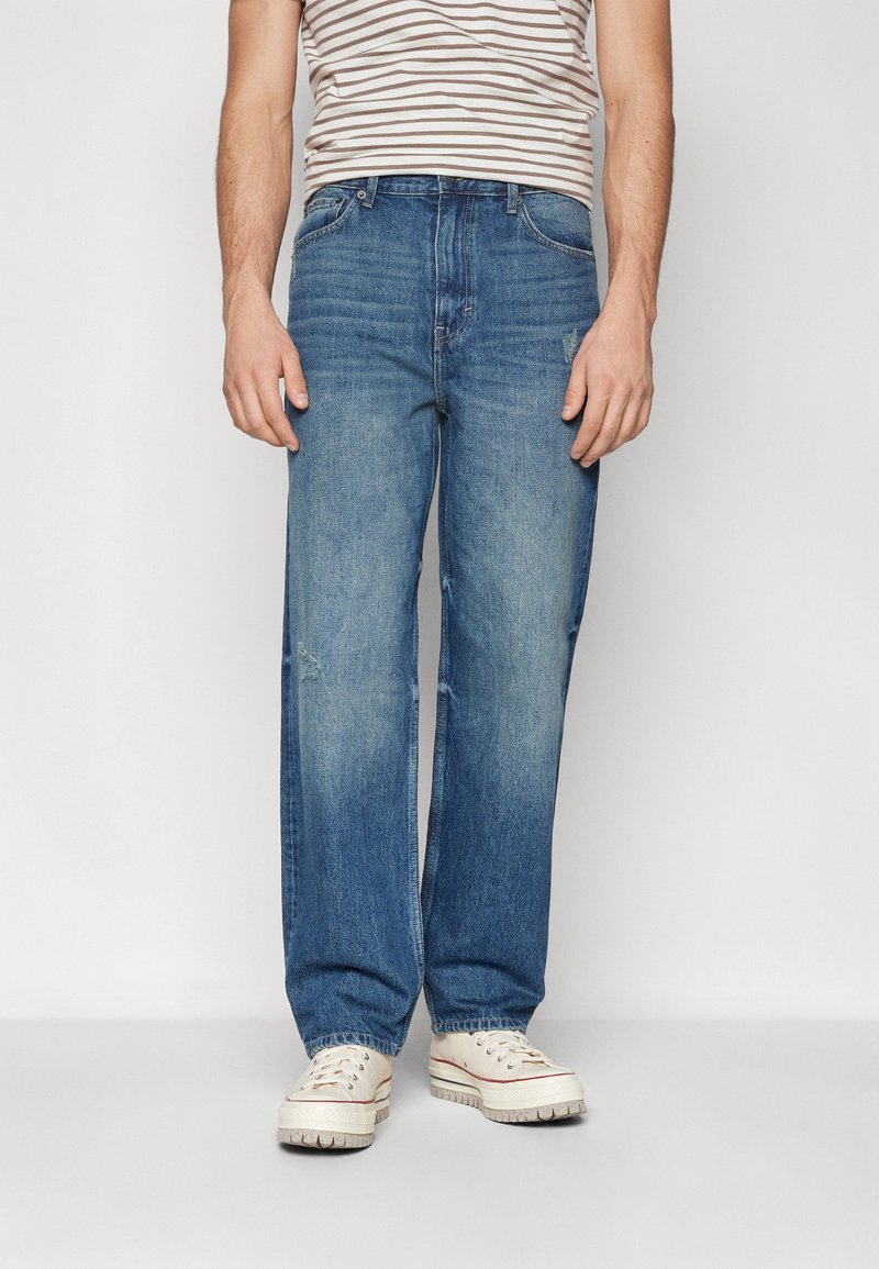 Jeans din denim de culoare albastru deschis, cu o croială lejeră, având zone decolorate și ușoare deteriorări. Asortate cu adidași de culoare crem și o cămașă în dungi.