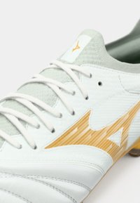 Λευκό ποδοσφαιρικό παπούτσι με υφή στην επιφάνεια, διαθέτει ανοιχτό γκρι λεπτομέρειες και πορτοκαλί διαγώνιες ρίγες. Τα κορδόνια είναι λευκά, με ένα λογότυπο στη γλώσσα.