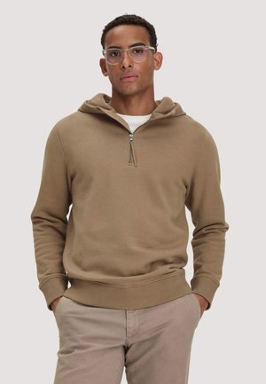 Man met kort krullend haar, die een bril met heldere glazen draagt, een lichtbruine half-rits hoodie en beige broek, staand met handen in de zakken.