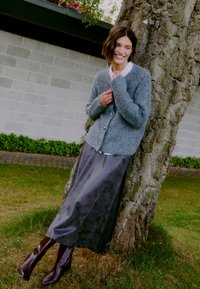 Gilet en tricot gris avec des boutons, associé à une longue jupe sombre. Bottes brunes à hauteur de genoux. Modèle appuyé contre un arbre en plein air.