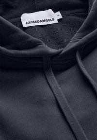Sweat à capuche gris foncé avec une texture douce, présentant une étiquette visible portant l'inscription "ARMEDANGELS" et un cordon de serrage au niveau de l'encolure.
