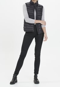 Černá prošívaná vesta s vysokým límcem, nošená přes světle šedé tričko s dlouhým rukávem, doplněná černými úzkými džíny a kotníkovými botami.