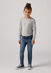 Mlada djevojčica s smeškom i smešnom smeškom, nosi smešnu majicu dugih rukava marki Levi's u sivoj boji, plave traperice i tamnoplave patike.