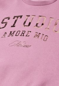 Tissu rose avec texte noir usé indiquant "STUDIO A MORE MIO" et texte cursif plus petit "Milano" en dessous.