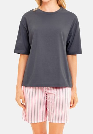 Person trägt ein lockeres dunkelgraues Kurzarmshirt und rosa-weiß vertikal gestreifte Shorts und steht vor einem einfarbigen Hintergrund.