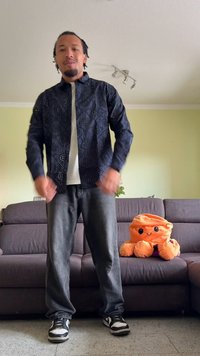 Casaco preto com lantejoulas e padrão riscado, camisa branca por baixo, jeans cinzentos e sapatilhas pretas, com um brinquedo de pelúcia laranja ao lado de um sofá.