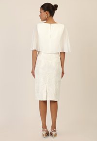 White dress featuring a textured floral pattern, knee-length, with a sheer cape-like overlay and a back zipper. Heeled sandals complete the look.