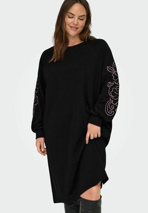 Kvinne iført en lang svart kjole med rosa blomsterbroderier på ermene, stående mot en ensfarget lys bakgrunn.