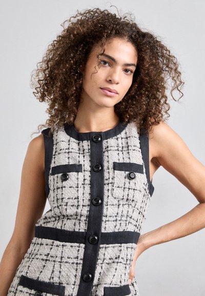 Robe sans manches, de longueur genou en tissu texturé noir et blanc avec un motif à carreaux. Dispose de boutons noirs et de deux poches poitrine.