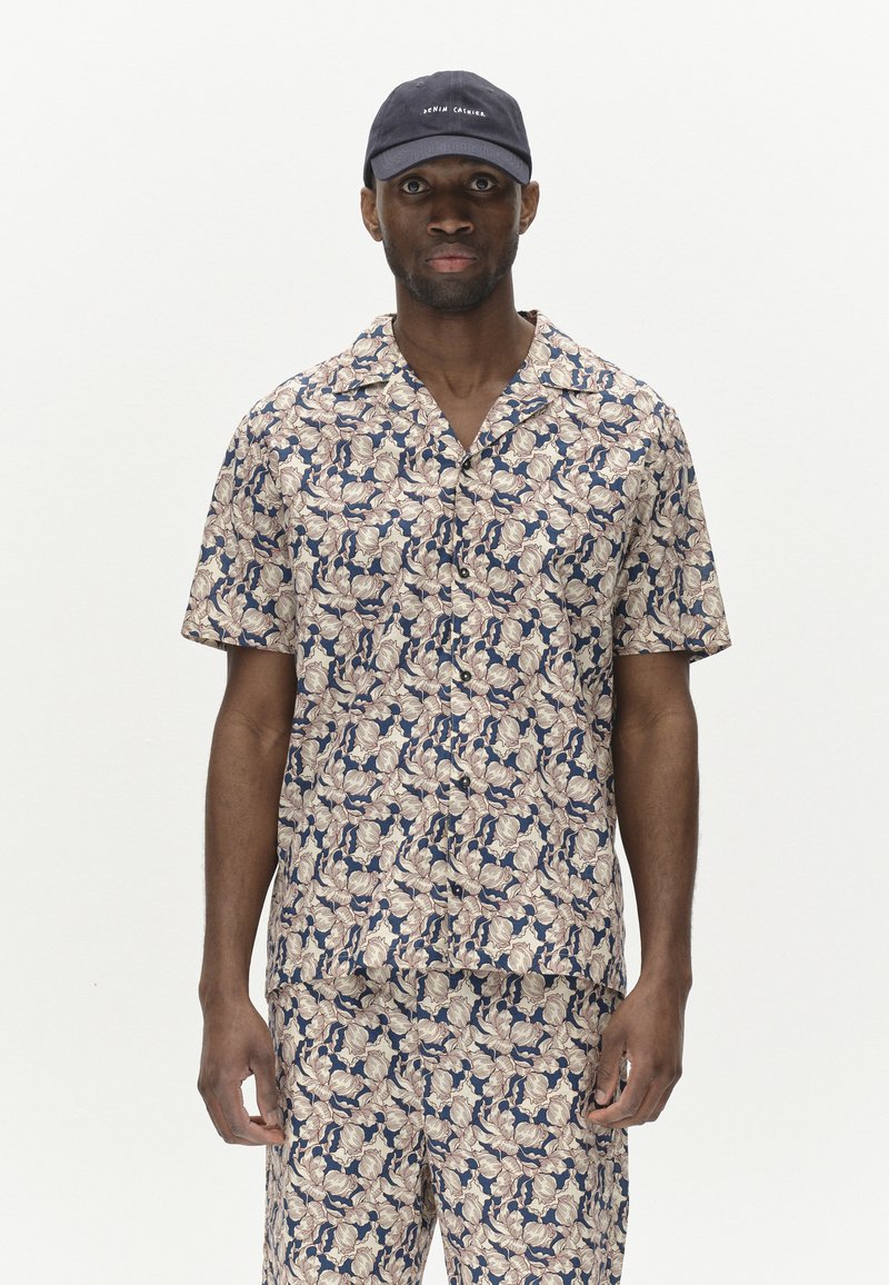 Camicia a maniche corte e pantaloni coordinati con un motivo floreale crema e blu navy, realizzati in tessuto leggero; abbinati a un cappellino blu scuro.
