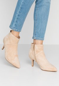 Botines de ante beige con puntera afilada, un sutil cierre lateral y un tacón de aguja de 7 cm, diseñados con un detalle de puño plegado.