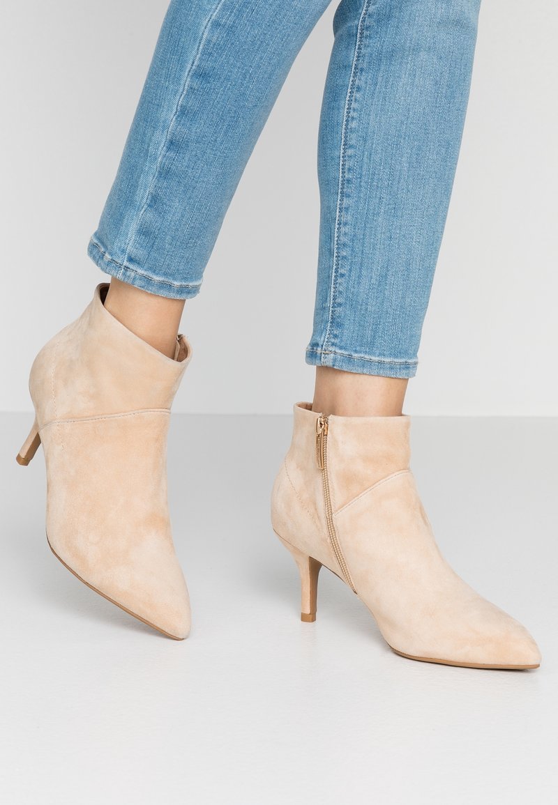 Botines de ante beige con puntera afilada, un sutil cierre lateral y un tacón de aguja de 7 cm, diseñados con un detalle de puño plegado.