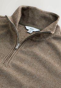 Brasls zip-up džemperis ar augstu apkakli, ar smalku suņu zobu rakstu un sudraba rāvējslēdzēju. Iekšpusē redzama birka ar zīmola nosaukumu.