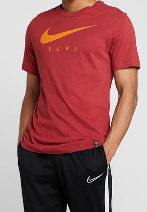 Homme portant un t-shirt Nike Roma rouge et un pantalon Nike noir, les mains dans les poches, sur un fond uni.