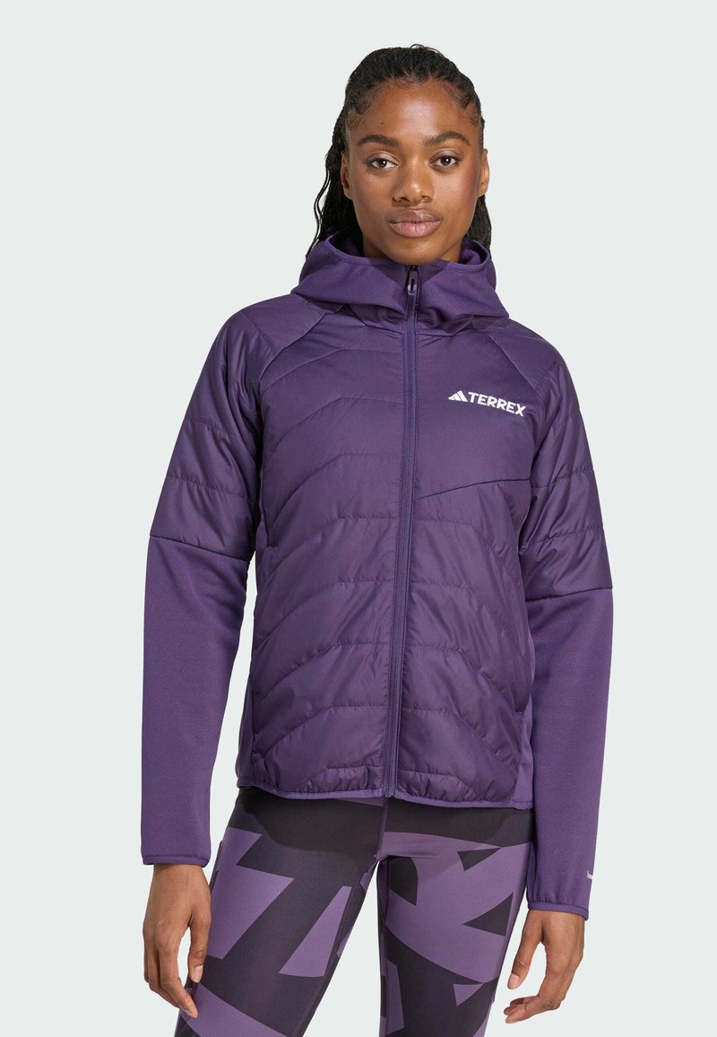 Kvinna i en lila Terrex-jacka och mönstrade leggings står mot en ljusgrå bakgrund och ser självsäkert in i kameran.