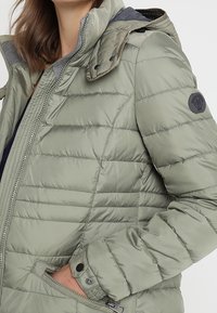 Chaqueta acolchada verde claro con cierre de cremallera, capucha desmontable y un parche en la manga. Presenta bolsillos laterales y puños elásticos.