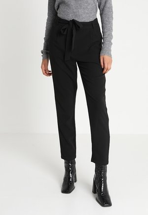 Femme portant un pantalon paperbag noir taille haute avec un nœud à l’avant, un pull gris côtelé et des bottines noires texturées à talons.