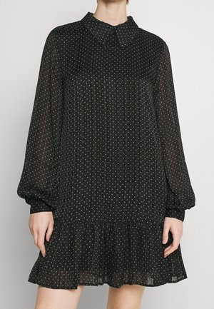 Woman wearing a black, long-sleeve, pleated mini dress with white polka dots and a small collar against a plain background.