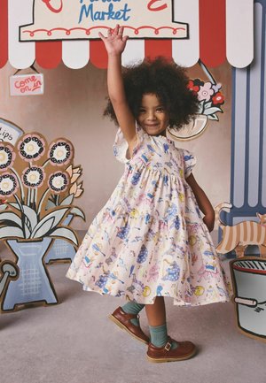 Smilende barn i en mønstret kjole løfter den ene armen, stående foran en fargerik, tegneserieaktig bakgrunn med blomster og marked.