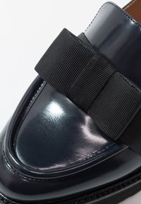 Mocassin en cuir verni marine avec une bande élastique noire texturée sur le devant ; finition brillante, bout arrondi et détail piqué le long de la semelle.