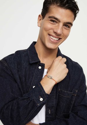 Bracelet nœud argenté sur cordon orange porté au poignet, associé à une chemise en denim foncé avec fermetures à boutons et finition texturée.