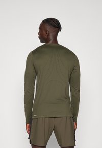 Olijfgroene sportshirt met lange mouwen van een gladde stof, voorzien van een geperforeerd ontwerp op de rug en een logo nabij de zoom.
