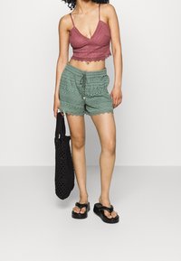 Kvinna som bär en mauve virkad midjetröja, gröna virkade shorts med dragsko, svarta platåsandaler och håller en svart virkad tote-väska.