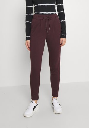 Frau trägt bordeauxfarbene, schmal zulaufende Hose mit Kordelzug am Bund, kombiniert mit weißen Sneakers und einem schwarz-weiß gestreiften Langarmshirt.