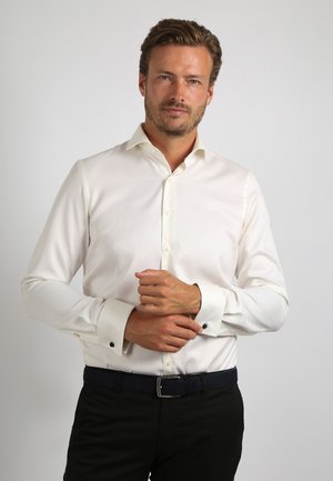 Witte langslijf shirt met een uitgespreide kraag, knoopsluiting en zwarte manchetten, gecombineerd met donkere broeken en een zwarte riem.
