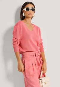 Helles rosa Strickpullover mit V-Ausschnitt, kombiniert mit passenden rosa Leinenhosen mit einem Gürtel in Schleifenoptik. Das Modell hält eine kleine cremefarbene Handtasche.