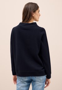 Mörkblå sweatshirt med hög krage, ribbade ärmslut och nederkant, visad bakifrån, tillsammans med ljusblå jeans. Slät textur.