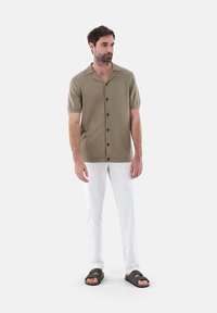 Camicetta khaki a maniche corte con chiusura a bottoni, abbinata a pantaloni bianchi e sandali scivolosi verde scuro. Tessuto liscio con una vestibilità rilassata.