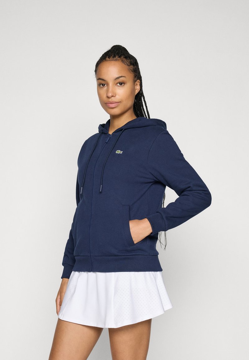 Marineblå zip-up hettegenser laget av myk stoff, med kengurulomme, snorhette og en liten grønn logo på venstre bryst.