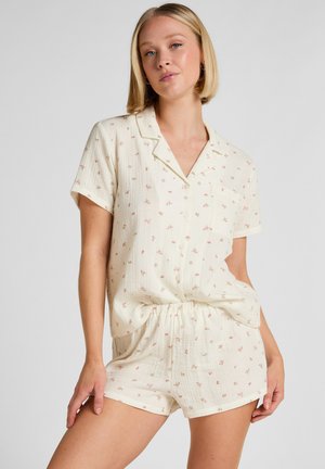 Vrouw draagt een crèmekleurige, korte mouwen blouse met knopen en bijpassende korte broek met kleine roze bloemenprint, poseert met hand op dij tegen een witte achtergrond.