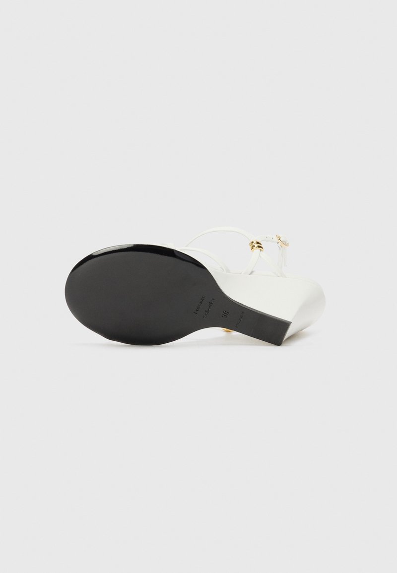 White heeled sandal featuring a black sole, minimalist strap design, and gold hardware accents. Smooth texture with a curved shape.