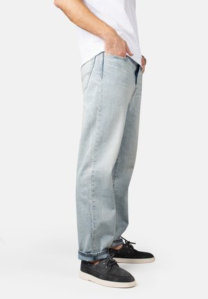 Hellblaue Denim-Jeans mit entspannter Passform, ausgestattet mit Gürtelschlaufen und Seitentaschen. Kombiniert mit dunklen Wildleder-Schuhen und einem weißen Hemd.